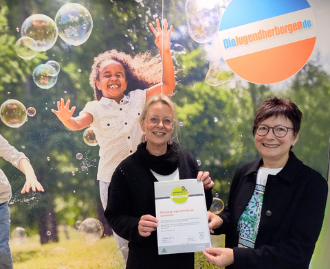 Zwei Frauen, die stolz ein Zertifikat halten, vor einer Fotowand mit Logo der Jugendherberge.