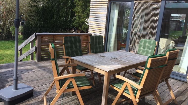 Ein einladender Terrassenbereich mit einem großen Holztisch und fünf Stühlen. Daneben steht ein Sonnenschirm und im Hintergrund sieht man einen Garten.