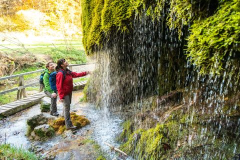 Zwei Wanderer stehen unter einem Wasserfall in einer grünen, bewaldeten Umgebung. Sie bewundern die Natur und genießen die frische Luft.