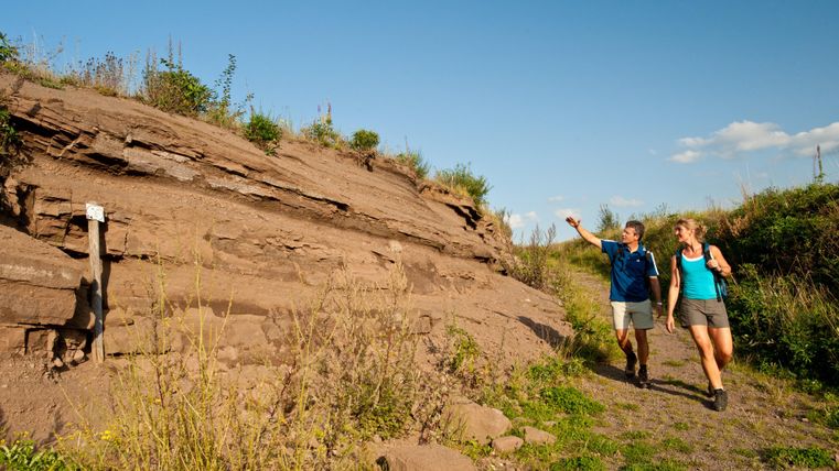 Zwei Wanderer bei Sommerwetter im Vulkangarten Steffeln. Einer der beiden zeigt auf die Felswand, an der sie vorbei wandern.