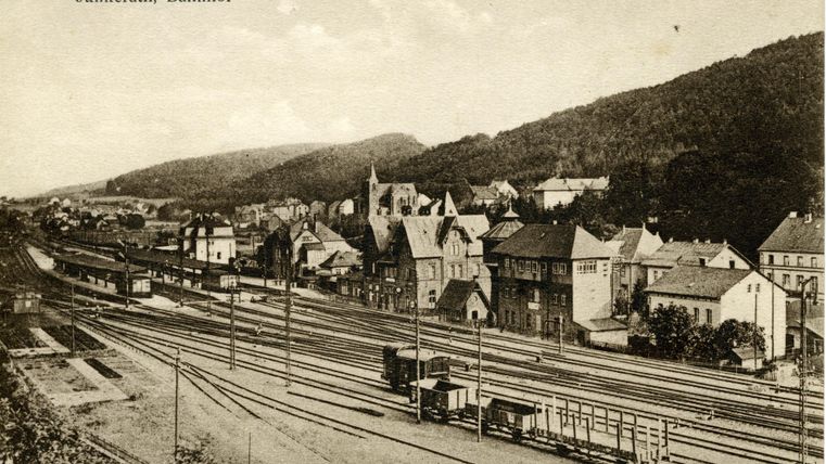 Historische Aufnahme des Bahnhofs Jünkerath mit mehreren Gleisen und umliegenden Gebäuden vor einer bewaldeten Hügellandschaft.