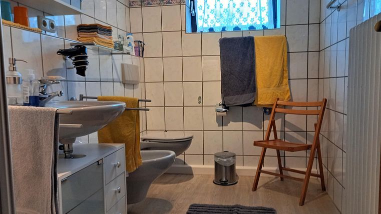 A modern bathroom with a sink, toilet, and a chair. The walls are tiled, and there are towels in various colors.