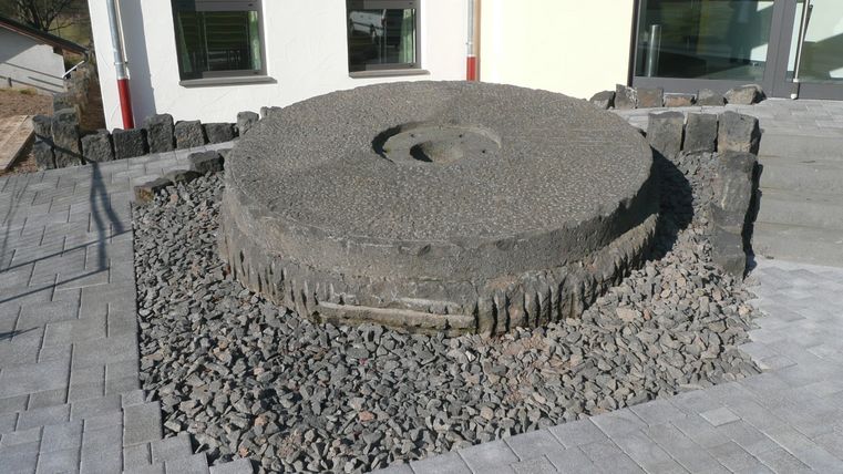 Großer Mühlstein auf einem Kiesbett vor einem Gebäude.