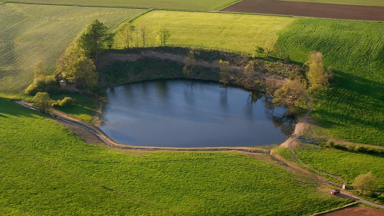 Un étang tranquille entouré de champs verts et d'arbres épars. Le paysage est vaste et accueillant.