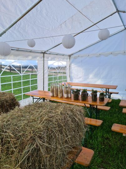 Une tente avec des tables en bois et des bancs, entourée de ballots de foin. Sur les tables, il y a des bouteilles en verre avec des éléments décoratifs.