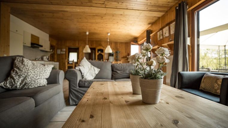 Ein gemütliches Wohnzimmer mit Holzwänden und einer großen Fensterfront. Auf einem Holztisch steht ein Blumenstrauß in einem Topf.