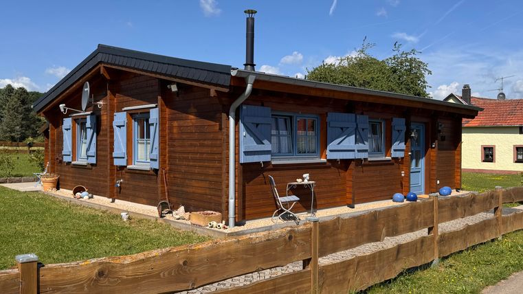 Ein gemütliches Holzhaus mit blauen Fensterläden. Es steht umgeben von einer grünen Wiese und einem niedrigen Holzzaun.