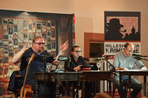 Zwei Männer und eine Frau sitzen nebeneinander an Tischen auf einer Bühne.