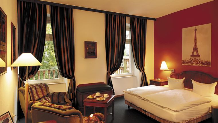 Une chambre d'hôtel confortable avec un lit double et un fauteuil. Les murs sont peints dans des couleurs chaudes et il y a des rideaux élégants ainsi qu'une image de la Tour Eiffel.