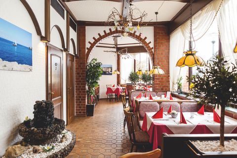 In einem Raum mit mediterranem Stil stehen mehrere Tische mit Stühlen und Sitzbänken in rot, der Boden ist befließt und im Raum stehen mehrere Pflanzen