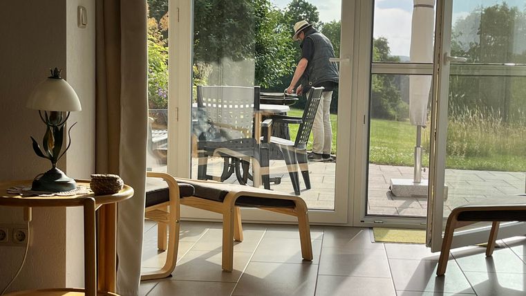 Un espace de vie confortable avec de grandes fenêtres donnant sur un jardin. À l'arrière-plan se trouve une personne qui travaille à une table.