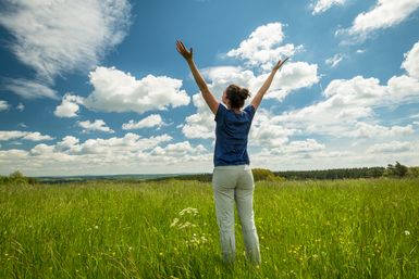 Eine Frau steht auf einer hochgewachsenen Wiese unter blauem Himmel, schaut in die Ferne und hält ihre Arm weit in die Luft.