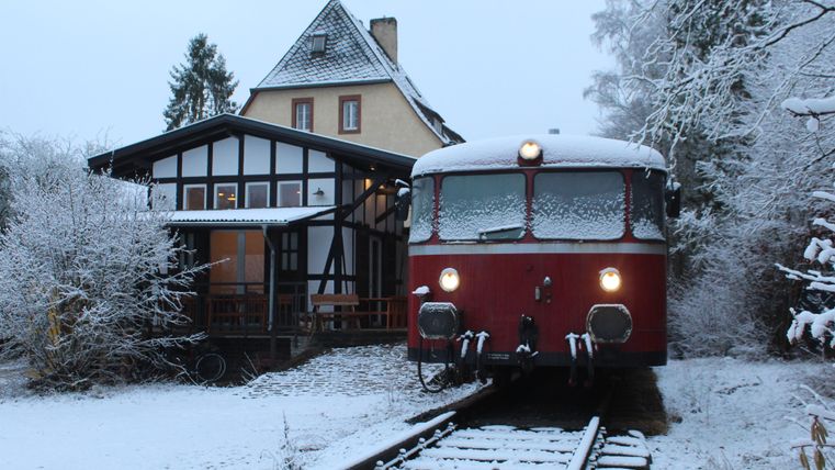 Ein roter, schneebedeckter Zug steht auf den Gleisen neben einem Gebäude in einer verschneiten Landschaft. Die Szene ist ruhig und friedlich, Schnee bedeckt den Boden und die Bäume.