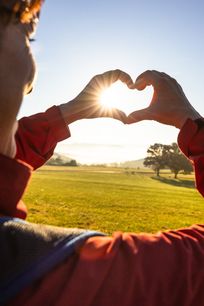 Eine Wanderin auf einer weitläufigen Wiese beim Sonnenaufgang zeigt ein Herz mit ihren Händen.