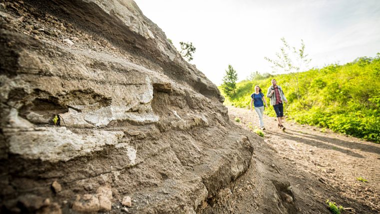 Zwei Personen wandern auf einem Pfad neben einer Felsformation aus Vulkangestein.