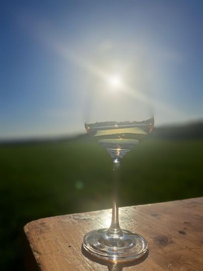 Een elegant wijnglas staat op een houten tafel. Op de achtergrond straalt de zon in een heldere blauwe lucht.