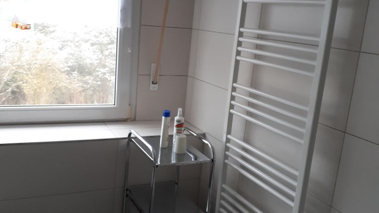 Ein helles Badezimmer mit einem Fenster und einem Handtuchheizkörper. Neben der Wand steht ein rollbarer Wäscheständer mit Pflegeprodukten.
