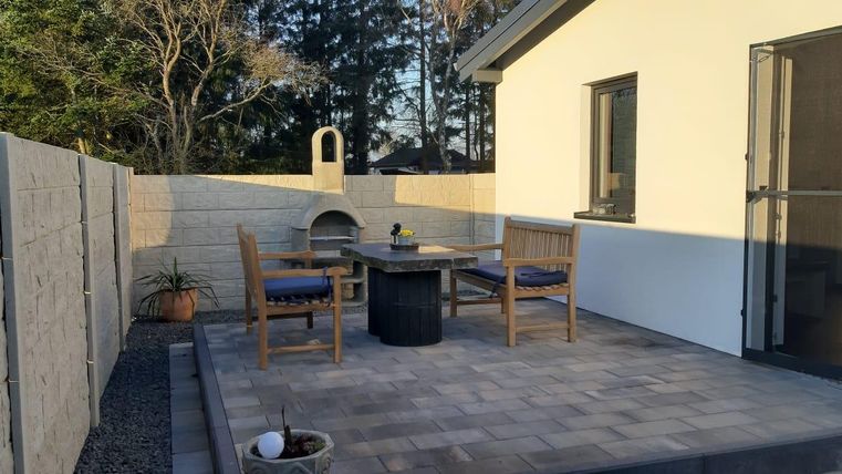 Eine gemütliche Terrasse mit einem Tisch und Stühlen aus Holz. Im Hintergrund sind Bäume und eine Wand aus Stein sichtbar.