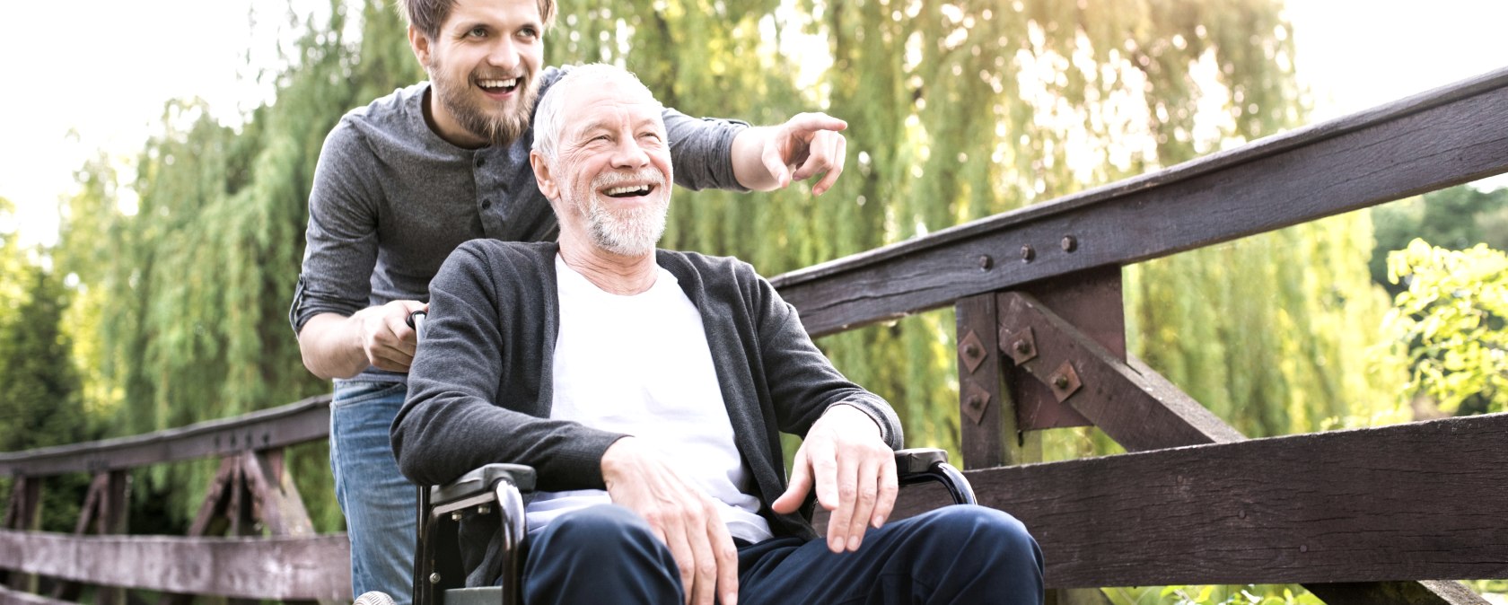 An elderly gentleman in a wheelchair is pushed over a bridge by a younger man. The two are in a good mood.