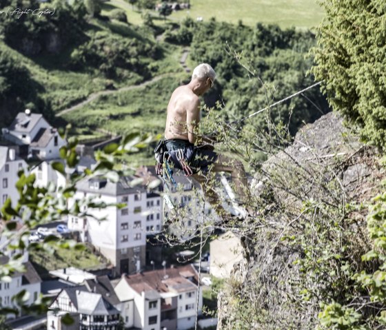 A shirtless man climbs a rock covered with plants, secured with ropes. Houses and green countryside can be seen in the background.
