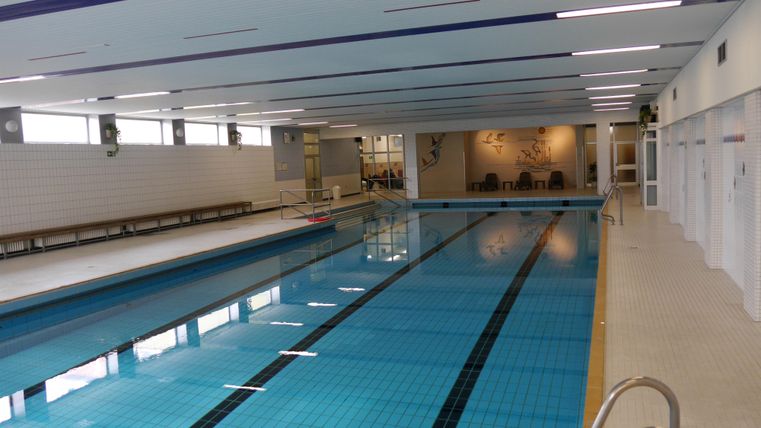 Innenansicht des Hallenbads mit einem langen Schwimmbecken, weißen Fliesenwänden und einem Sitzbereich im Hintergrund.