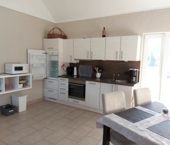 Einen schwarz-wei&szlig;e K&uuml;che steht an einer Wand, daneben Regale und ein gro&szlig;er Esstisch mit mehreren St&uuml;hlen.