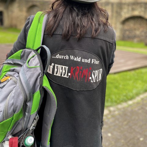 Eine Hut tragende Person steht mit dem R&uuml;cken zum Betrachter vor einer gro&szlig;en Stadtmauer. Sie tr&auml;gt eine Jacke mit der Aufschrift "...durch Wald und Flur auf Eifel-Krimi-Spur" auf dem R&uuml;cken und tr&auml;gt &uuml;ber einen Schulter einen Rucksack.