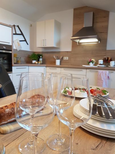 Eine moderne Küche mit hellen Möbeln und Holzelementen. Auf dem Tisch stehen Weingläser sowie frische Speisen angerichtet.