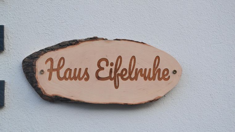 Een houten bord met de inscriptie "Haus Eifelruhe". Het is aan een witte muur bevestigd.