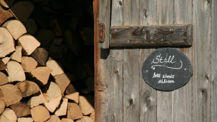 Een houten schuur met een bord waarop "Stal" staat. Naast de deur ligt een stapel met opgestapeld hout.