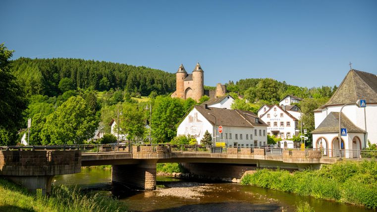 Die Bertradaburg thront über einem Dorf in der Eifel, umgeben von grünen Wäldern. Im Vordergrund fließt die Kyll unter einer Brücke hindurch.