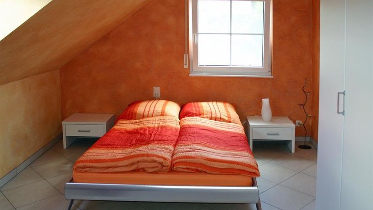 Een gezellige slaapkamer onder het dak met een tweepersoonsbed en oranje dekens. Naast het bed staan twee nachtkastjes en een groot raam zorgt voor natuurlijk licht.