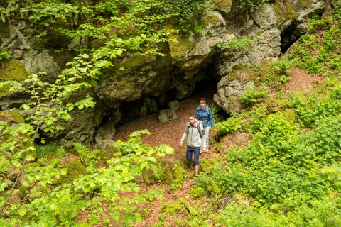 Zwei Wanderer kommen aus einem Höhleneingang einer massiven Felswand hinaus in einen dichten Laubwald.