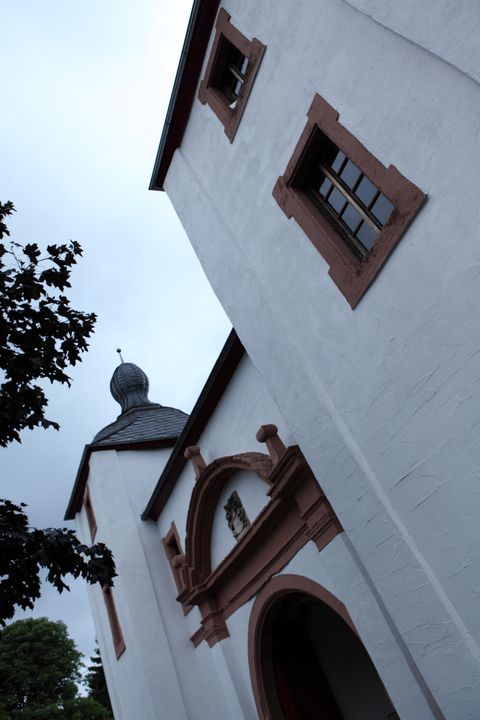 Weißes Gebäude mit roten Fensterrahmen und einem Turm mit geschwungenem Dach. Der Himmel ist bewölkt, ein Baum ist im Vordergrund sichtbar.