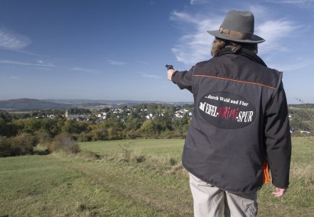 Person mit Hut und Jacke mit Aufschrift 'auf EIFEL KRIMI SPUR' zeigt auf eine weite Landschaft mit einem Dorf im Hintergrund., © Kappest