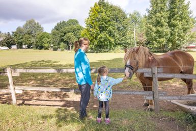 Eine Frau und ein Kind stehen an einem Holzzaun, hinter dem ein Pferd steht. Das Kind streichelt das Pferd.