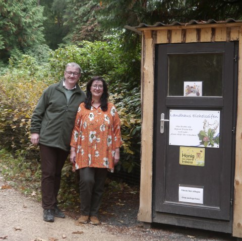 Ein Mann und eine Frau stehen neben einem kleinen Holzhaus mit Aufschrift Landhaus Eichelseifen.