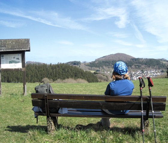 Eine Person sitzt auf einer Bank und blickt auf den Nerother Kopf. Im Vordergrund sind Wanderstöcke und ein Rucksack zu sehen., © Touristik GmbH Gerolsteiner Land, Ute Klinkhammer