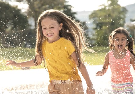 Two girls run through a pool of water one after the other, laughing, and the water splashes in all directions.
