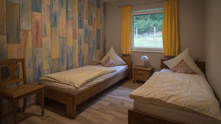 A simple bedroom with two single beds and a colorful wood design on the wall. The window lets in plenty of daylight and there are yellow curtains.