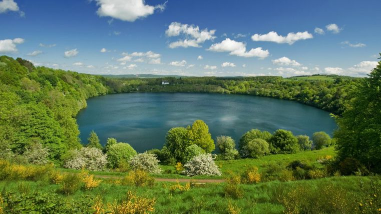 Un lac pittoresque entouré d'une verdure luxuriante et de fleurs colorées. Le ciel est clair avec quelques nuages.