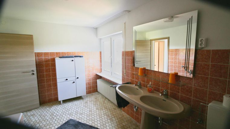 Ein helles Badezimmer mit orangefarbenen Fliesen und einem großen Spiegel. Es enthält ein Waschbecken, eine Toilette und einen weißen Schrank.