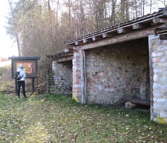 A person looks at an information board next to a historic lime kiln made of stone in Nohn. The kiln stands at the edge of the forest., © Touristik GmbH Gerolsteiner Land, Ute Klinkhammer