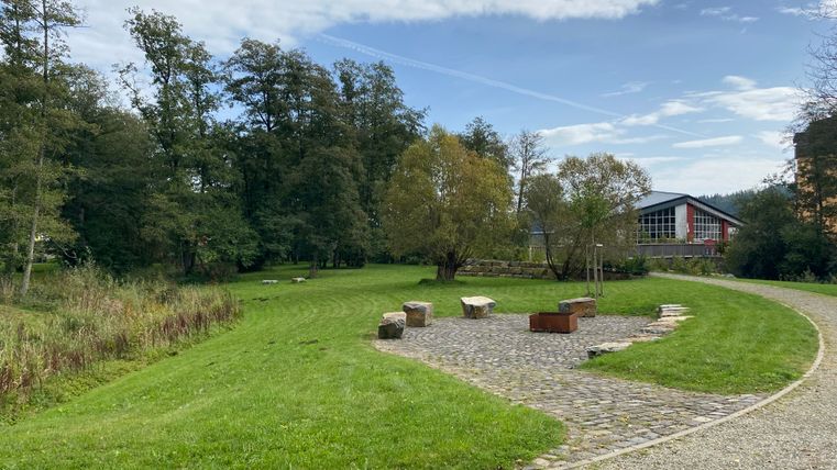 Barrierefreier Spazierweg durch den Kurpark Stadtkyll mit Grillschale und Steinen als Sitzgelegenheit. Gebäude im Hintergrund.
