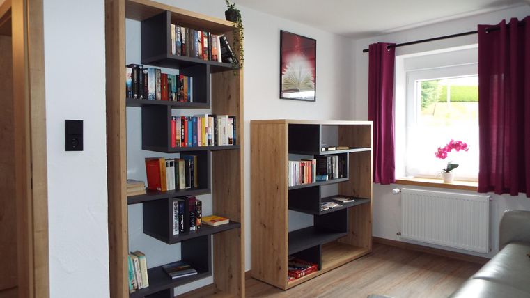 Ein gemütliches Wohnzimmer mit zwei Bücherregalen und einem Fenster. Die Wände sind hell, und rote Vorhänge bringen Farbe in den Raum.