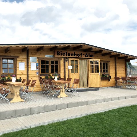 Holzgeb&auml;ude mit Terrasse und mehreren Tischen und St&uuml;hlen davor. 