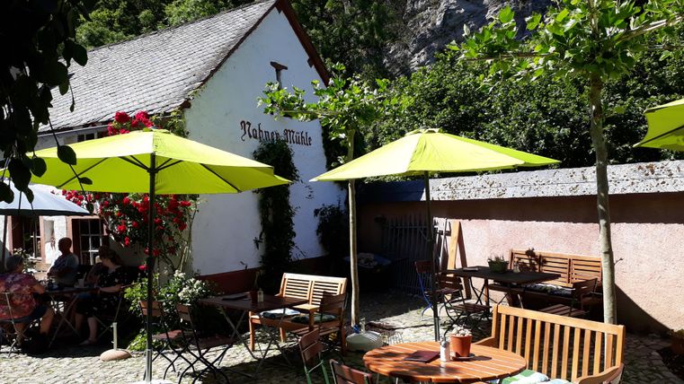 Gemütlicher Außenbereich der Nohner Mühle mit grünen Sonnenschirmen, Holzmöbeln und blühenden Rosen an der Wand. Menschen sitzen entspannt im Schatten.