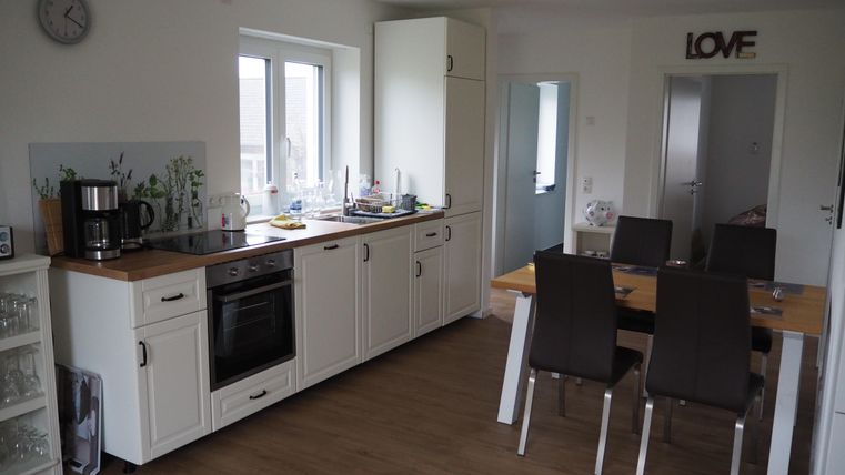 Eine moderne Küche mit weißen Schränken und Holzoberflächen. Ein Esstisch mit Stühlen ist ebenfalls im Raum.