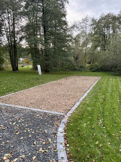 Ein leerer Platz mit Sand, umgeben von Grünflächen und Bäumen. Im Hintergrund steht ein Schild.