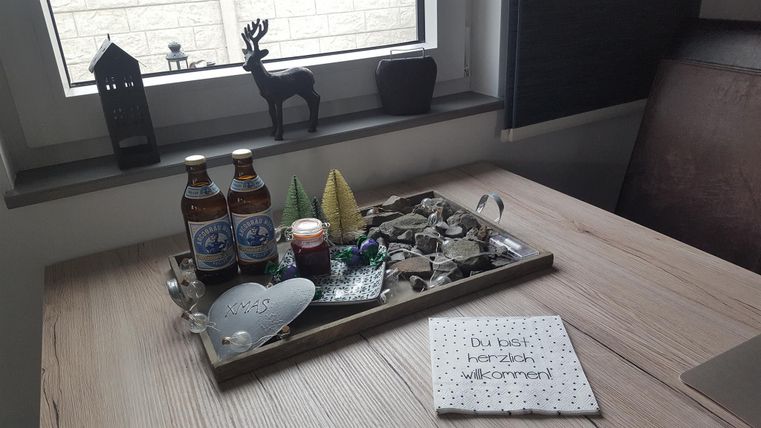 Ein dekorativer Tisch mit einer Holzplatte, auf dem sich zwei Flaschen Bier, ein kleiner Kerzenhalter und verschiedene dekorative Elemente befinden. Ein Schild mit der Aufschrift „Du bist herzlich willkommen“ liegt ebenfalls auf dem Tisch.